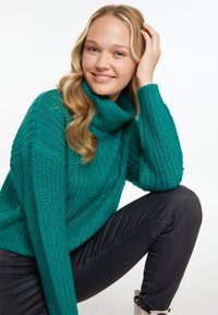 Pulover tricotat de culoare verde-albastrui, cu guler înalt și model texturat, asortat cu blugi negri. Modelul stă în genunchi, având o mână în păr.