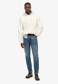 Hvid hættebluse med prægede logo, lagdelt over en lys blå ternet skjorte. Båret med blå jeans og mørkebrune snøresko.
