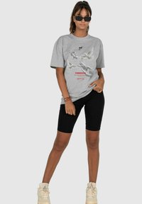 Graues Baumwoll-T-Shirt mit Grafikdruck, der Vögel und das Wort "FREIHEIT" in Rot zeigt. Kombiniert mit schwarzen Radshorts und hellen Sneakers.
