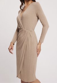 Femme portant une robe portefeuille beige à manches longues avec un devant drapé et un ourlet arrivant au genou, debout devant un fond blanc uni.