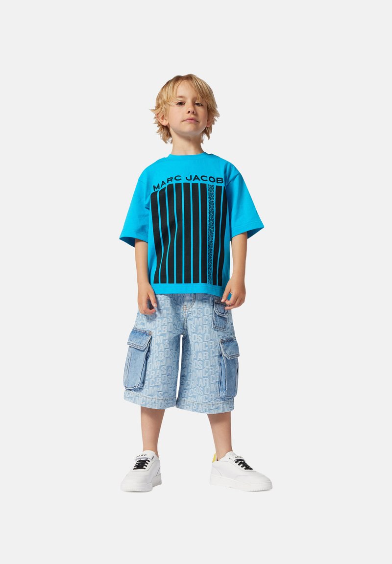 Maglietta grafica blu con strisce verticali nere, abbinata a pantaloni cargo azzurri chiari con loghi ripetuti. Sneakers bianche completano il look.