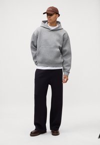 Szara,oversize'owa bluza z kapturem z przednią kieszenią, w połączeniu z luźnymi granatowymi spodniami. Brązowa czapka i ciemnobrązowe buty dopełniają strój.