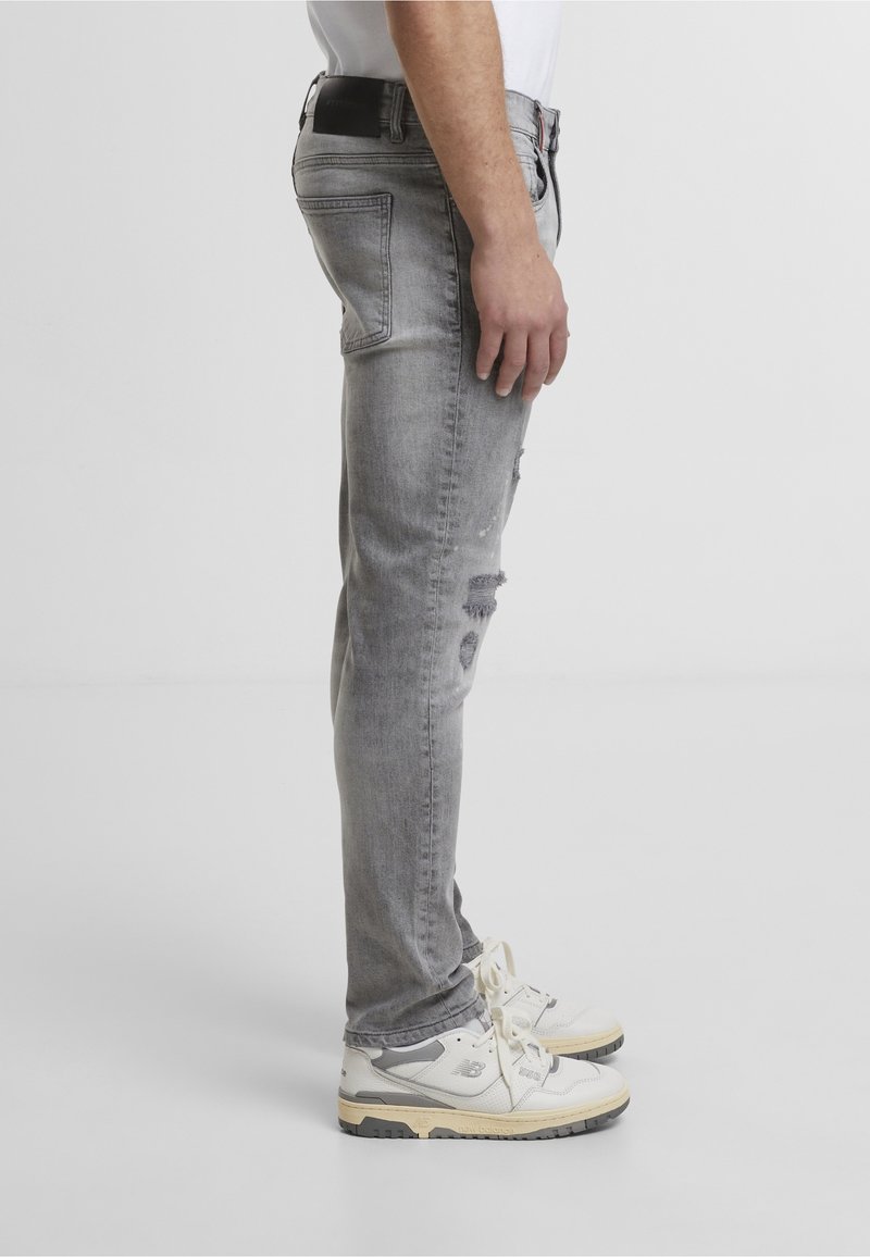Homme debout en jean gris déchiré et baskets blanches avec semelles noires sur un fond blanc uni, vue de côté.
