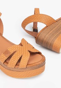 Sandalias de cuña en tono tostado con tiras trenzadas y parte superior texturizada. La plataforma presenta estrías horizontales. Plantilla suave para mayor comodidad.