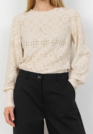 Femme portant un chemisier en dentelle crème à manches longues avec des motifs géométriques et un pantalon noir taille haute à fermeture par boutons.