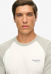 Biała koszulka z szarymi raglanowymi rękawami, z małym niebieskim logo "SuperDry Athletics" na piersi. Gładki materiał bawełniany.