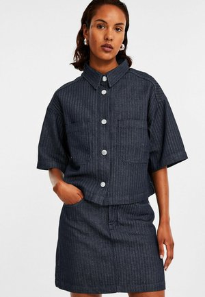 Donkerblauw denim shirt met korte mouwen, horizontale strepen, twee voorkruiden en zilveren knopen. Gecombineerd met een bijpassende denim rok.
