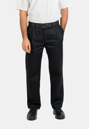 Man wearing black pleated trousers, black belt, black shoes, and a white short-sleeve shirt, standing with one hand in pocket.