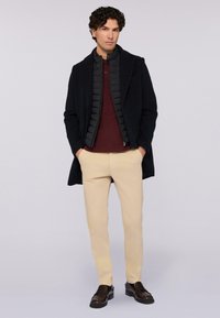 Cappotto nero con uno strato interno a effetto trapuntato sopra un polo bordeaux, abbinato a chinos beige e mocassini marroni.