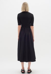 Schwarzes Kleid mit einem enganliegenden, gerippten Oberteil, kurzen Ärmeln und einem fließenden, gerafften Rock. Getragen mit flachen schwarzen Sandalen. Das strukturierte Material scheint weich zu sein.