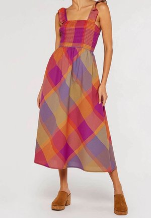 Kobieta w bezrękawnikowej, wielokolorowej sukience midi w kratę z falbankami na ramiączkach i brązowych chodakach na platformie, stojąca na jednolitym tle.