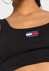 Ingrandimento di una persona che indossa un crop top nero di Tommy Jeans con un logo rettangolare ricamato in bianco, rosso e blu navy sul petto.
