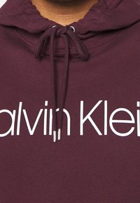 Sudadera burdeos con capucha ajustable, que presenta un logo blanco de "Calvin Klein" en el pecho y detalles de canalé en el cuello.