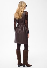 Blondynka w brązowej sukience z materiału przypominającego skórę, przewiązanej w talii, na przezroczystej, kwiecistej bluzce z długim rękawem, czarnych legginsach i brązowych, zamszowych kozakach do kolan, stojąca.