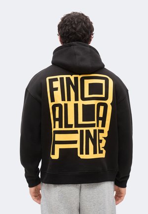 Un homme portant un sweat à capuche noir avec de grandes lettres jaunes en bloc indiquant "FINO ALLA FINE" dans le dos, debout devant un fond uni.