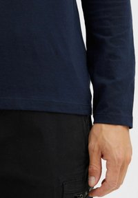 Navy long-sleeve T-shirt made of soft cotton, featuring a rounded hem and simple stitching; worn with black pants showing a pocket detail.