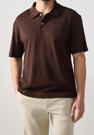 Bruine poloshirt met een zachte textuur, knoopkraag en een borstzak. Gedragen met lichtgekleurde broek voor contrast.