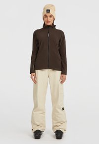 Fleece bunda tmavě hnědé barvy s vysokým límcem a celopropínacím zipem, v kombinaci s světle béžovými cargo kalhotami a krémovou čepicí. Viditelné černé boty.