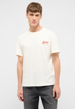 Junger Mann trägt ein weißes T-Shirt mit kurzen Ärmeln und einem kleinen roten "Jeans Company Mustang"-Logo auf der linken Brustseite und dunkle Jeans.