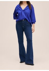 Haut en satin bleu à col en V et coupe ample ; assorti à un jean évasé en denim foncé. Bottes visibles en bas. Texture lisse.