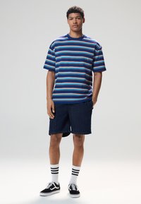 Stripet T-shirt i blå, rød og gul; afslappet pasform. Kombineret med mørke denimshorts og sorte sneakers, med hvide sokker med striber.