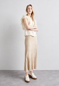 Beige blus med volanger och korta ärmar, tillsammans med en ribbad maxikjol och vita plattformsskor. Neutrala färger och texturer dominerar.