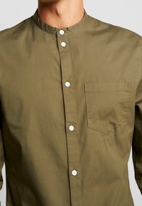 Man i olivgröna skjortor med mandarin-krage, bröstficka och vita knappar i mitten.