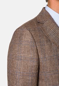 Blazer en tweed marron avec un motif à carreaux bleu et beige porté sur une chemise rayée bleue et blanche.