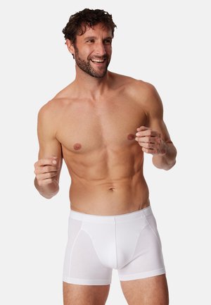 Boxer in cotone bianco con design senza cuciture e vita elastica. Il tessuto sembra morbido e leggero. Il modello è in piedi, sorridente.