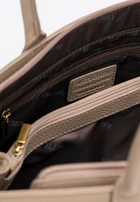 Intérieur de sac à main texturé beige avec plusieurs compartiments, présentant un doublure en tissu foncé et une étiquette. Accents de fermeture éclair dorés visibles.