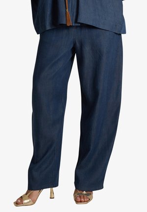 Pantaloni blu scuro a gamba dritta indossati con sandali dorati con tacco alto e punta aperta che mostrano le unghie dei piedi smaltate.