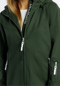 Veste verte légère avec capuche. Comprend des zips blancs, un logo sur le col intérieur et plusieurs poches zippées. Texture lisse.