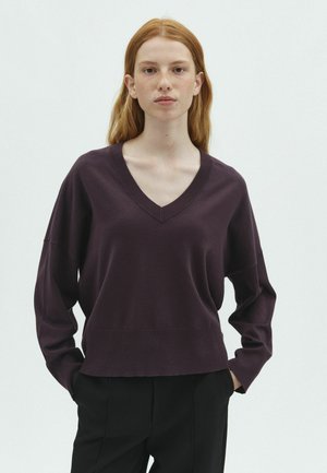 Jeune personne aux longs cheveux roux portant un pull violet foncé à col en V et un pantalon noir, debout les mains dans les poches devant un fond uni.