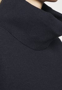 Laivastonsininen poolopaita, jossa on pehmeä, teksturoitu kangas ja leveä, taitettu kaulus. Minimalistinen estetiikka, jossa on puhtaat linjat.