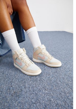 Baskets montantes Nike avec des accents bleu clair, beige et rose, présentant des orteils perforés et des lacets blancs, portées avec des chaussettes blanches côtelées.