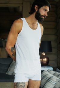 Hvid tanktop og trusser på en mand med skæg, der viser en tatovering på sin arm. Neutral baggrund med en lampe og ternede puder.
