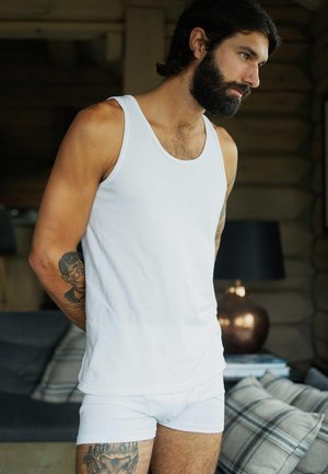 Hvid tanktop og trusser på en mand med skæg, der viser en tatovering på sin arm. Neutral baggrund med en lampe og ternede puder.