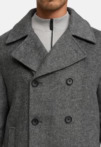 Cappotto grigio in lana a doppio petto con motivo a spina di pesce, ampio colletto a revers e bottoni neri. Include uno strato interno con zip.