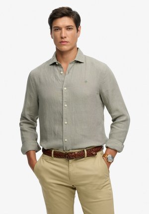 Jeune homme portant une chemise en lin gris clair boutonnée, manches retroussées, pantalon beige, ceinture tressée marron et montre-bracelet en or, les mains dans les poches.