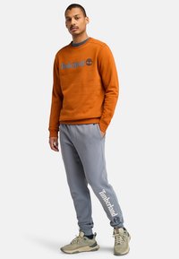 Orangener Sweatshirt mit "Timberland"-Logo, kombiniert mit grauen Jogginghosen, die einen Logo-Druck am Bein aufweisen, und hellen Schuhen. Lässiges Design.