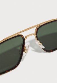 Ochelari de soare pătrați cu lentile de culoare verde închis, ramă din tortoise și accente din metal aurit. Dotati cu garnituri de nas clare pentru confort.
