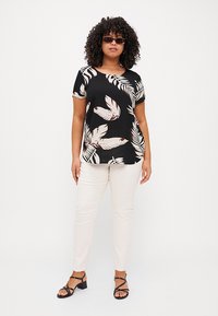 Schwarzes kurzärmliges Oberteil mit weißen und beige Blattmustern, kombiniert mit beigen Slim-Fit-Hosen und schwarzen Absatzsandalen.