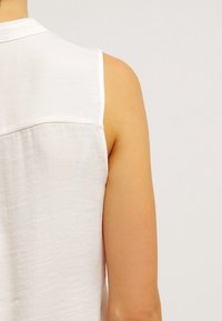 Blusa blanca sin mangas con textura suave, presenta una costura sutil en el canesú trasero y un escote redondeado.