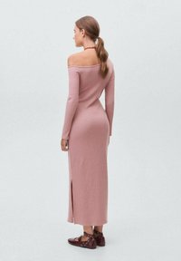 Ribbed, off-the-shoulder pink dress with long sleeves and side slit, paired with dark flat shoes featuring circular cutouts.