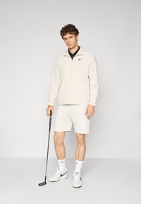 Men's golf apparel features a light beige zip-up jacket, black polo shirt, beige shorts, white socks, and white sneakers with black accents.