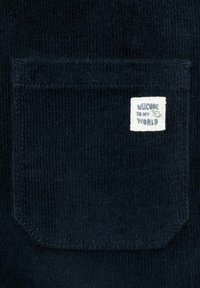 Tessuto in corduroy blu navy con una tasca quadrata e un'etichetta bianca che recita "BENVENUTO NEL MIO MONDO" con un piccolo grafico verde.