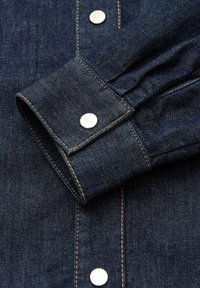 Dunkelblauer Denim-Hemdsärmel mit weißen Metall-Druckknöpfen, sichtbaren Nähten und einer strukturierten Stoffoberfläche. Der Ärmelbund weist leichte Falten auf.