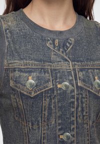 Gros plan d'une personne portant un gilet en denim sans manches avec des boutons en métal et des coutures orange sur une chemise grise côtelée.