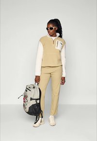 Frau in beiger Fleecejacke, passender Hose, weißen Turnschuhen, Sonnenbrille, mit einem grau-schwarzen Rucksack vor einem einfarbigen Hintergrund.