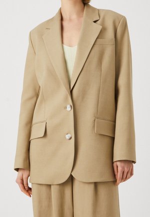 Beige op maat gemaakte blazer met notch-kleppen, twee knopen en zijzakken. Gladde textuur en gestructureerde pasvorm. Draag een lichtgroene top eronder.